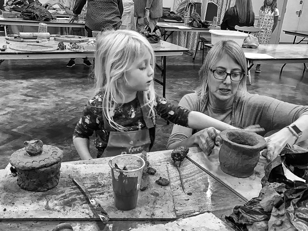 Woman crouching to help a little girl standing behind a table make a clay pot.