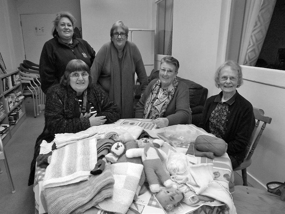 Three women sitting and two standing behind a table displaying knitted crafts including towels and a teddy bear.