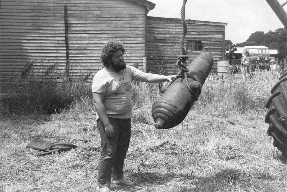 Black-and-white photo of a man steadying a ship's cannon being lifted by a crane.