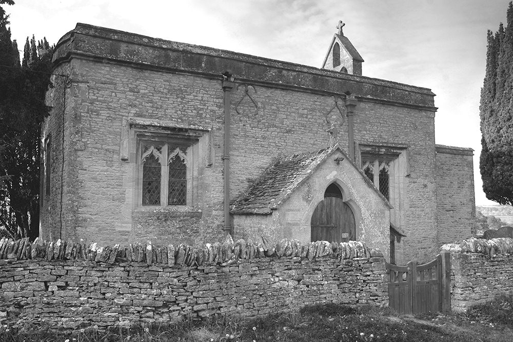 Small stone church, with no steeple, quite box-like in shape with stained glass windows and a porch.