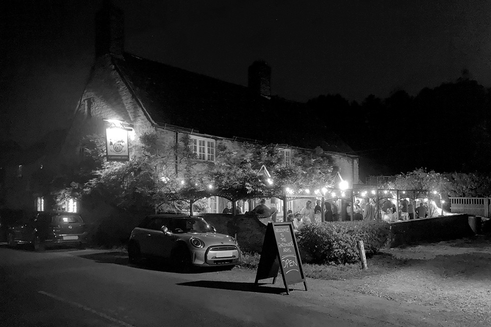 View of the pub from the road, lit up at night, with lots of people in the garden.