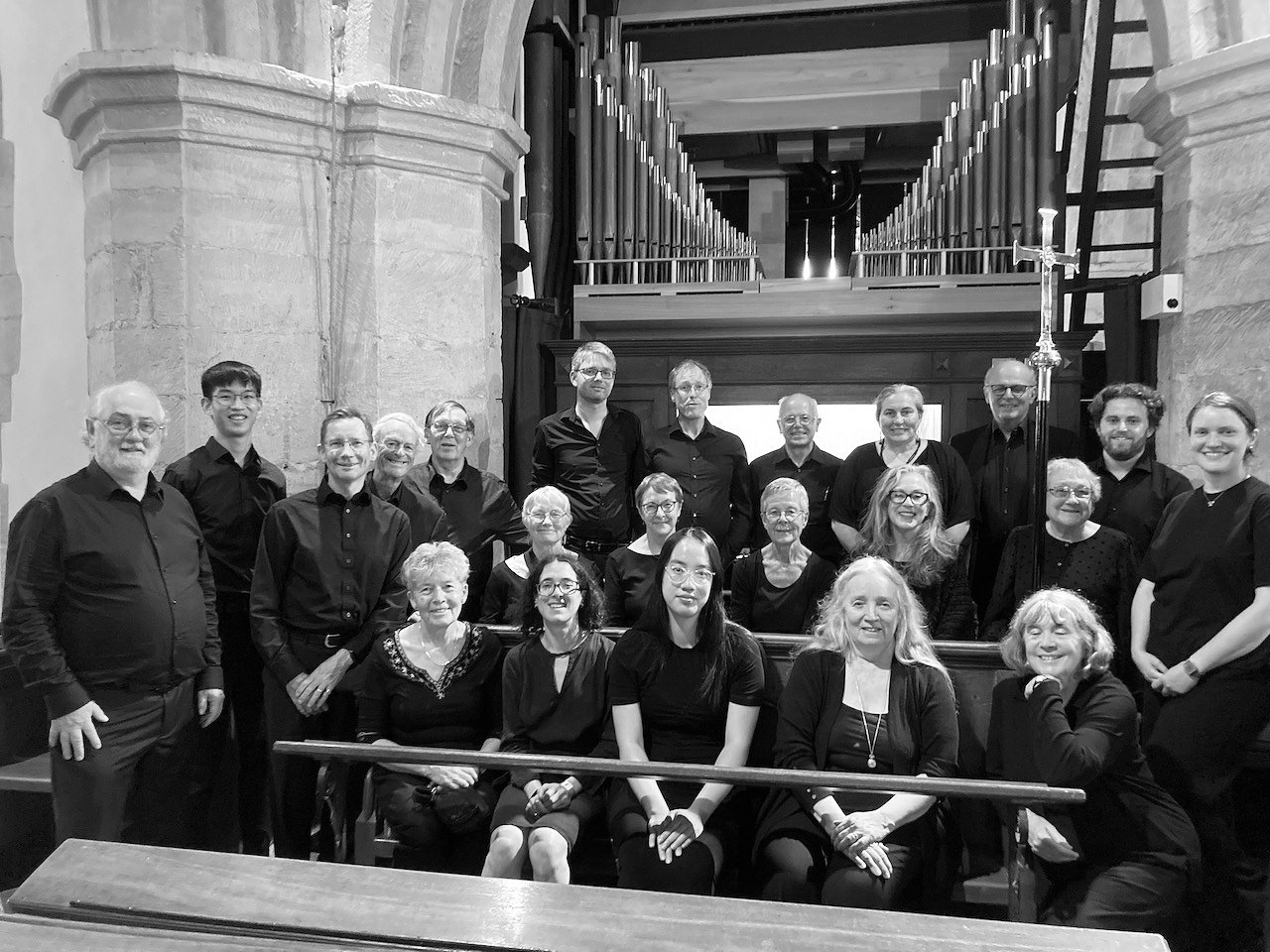 About 20 choir singers sitting on pews in front of a pipe organ, smiling for the camera.