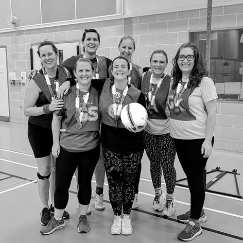 Group photo in a sports hall. Seven women wear netball bibs and medals, smiling. One holds a ball.